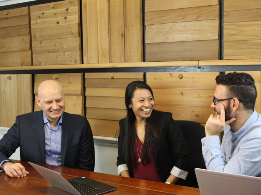 Colleagues smiling during a meeting around a table with laptops in an office with wooden decor.